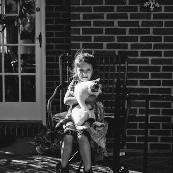 Black and white documentary portrait of young girl holding calico cat in her lap in a rocking chair in front of a brick house on a porch with partial woman on left side and partial figure on right side framing her 