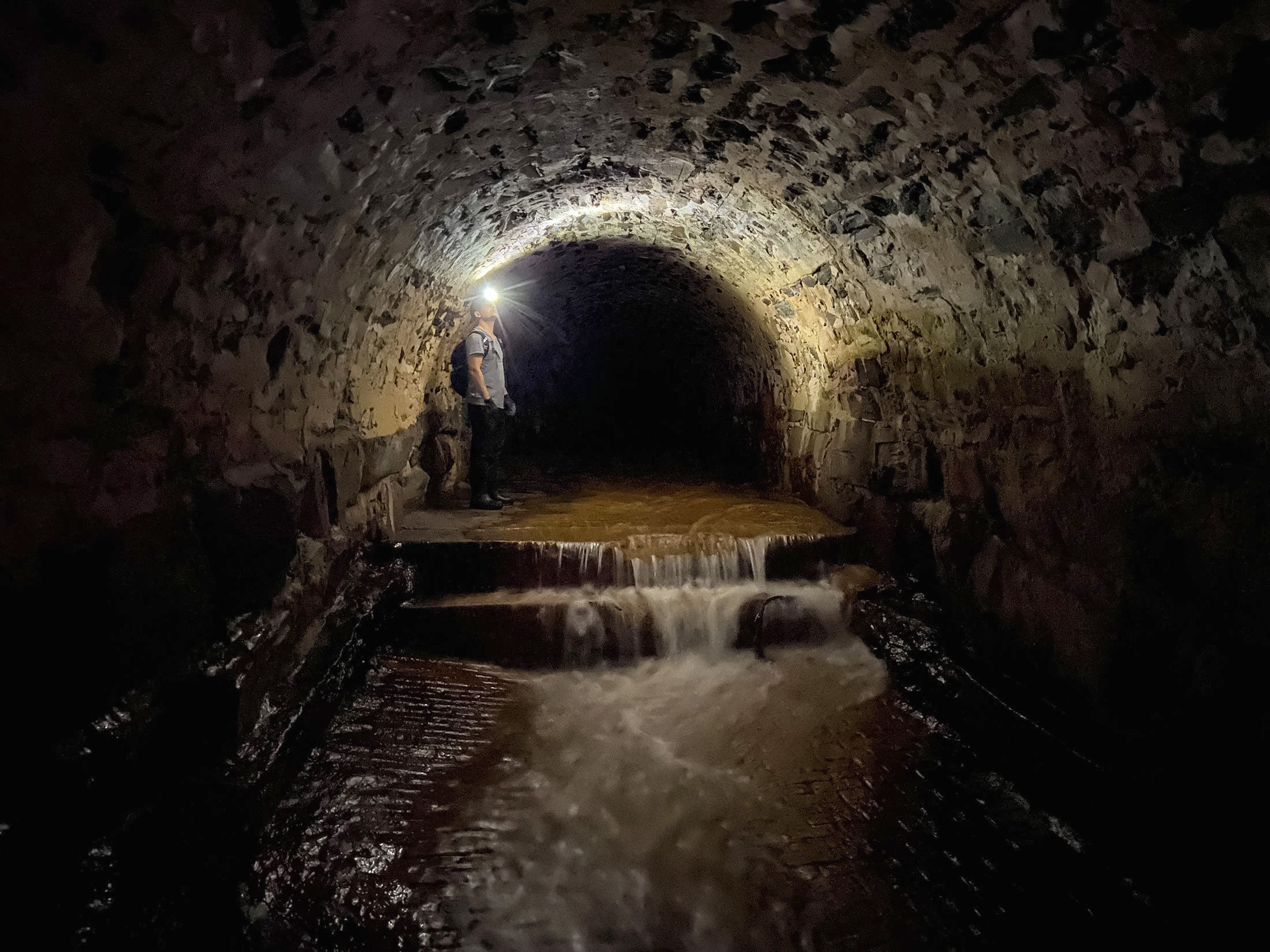 Photos of a man with a headlamp and waders standing in an underground tunnel with an arched stone ceiling. Water cascades through the tunnel down a set of steps.