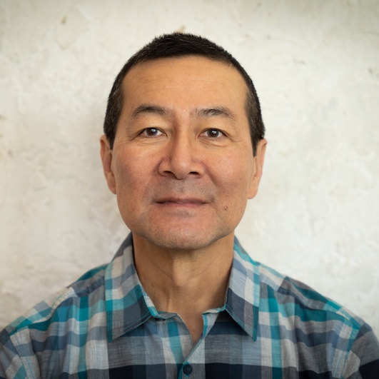 Don Lee: Man with short dark hair wearing a teal and gray plaid shirt, photographed against a textured light wall