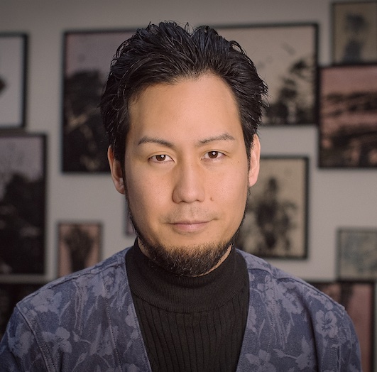Kei Ito: Man with dark swept-back hair and a goatee, wearing a black turtleneck and a blue floral-patterned jacket, photographed in front of a wall hung with framed photographs.