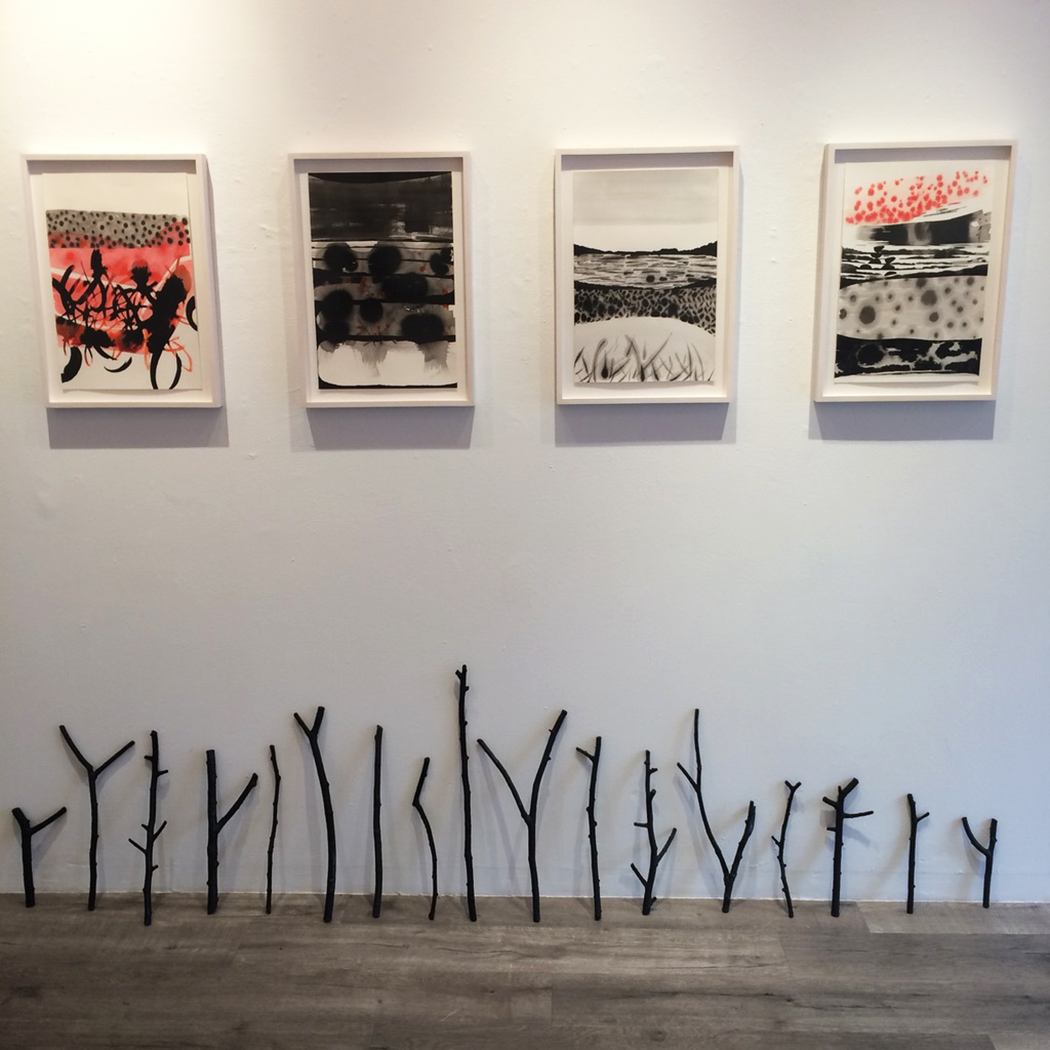 Installation view of framed drawings by Magnolia Laurie and black stick sculptures leaning against a wall. 