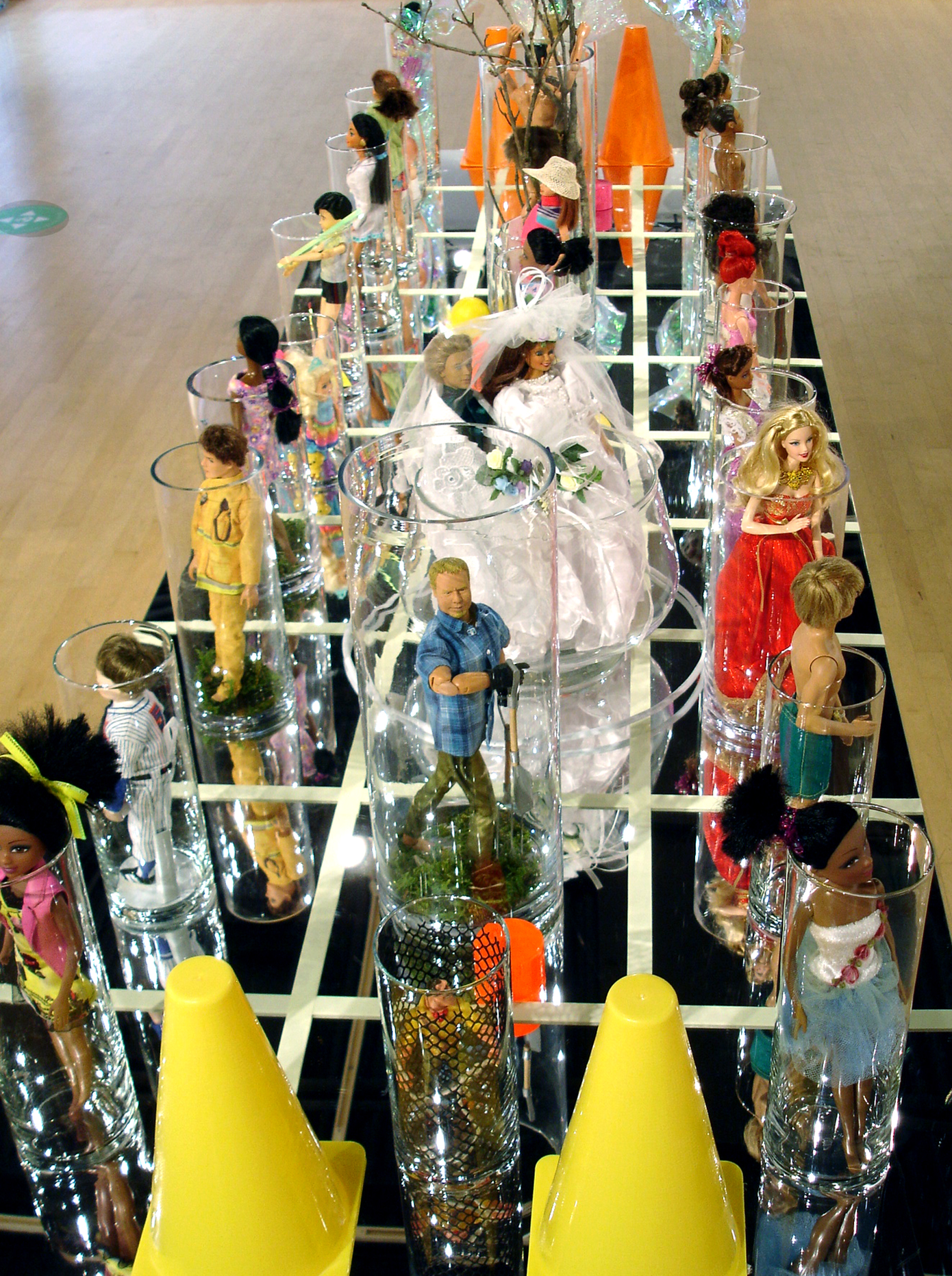 "A New Playing Field" is an installation staging of "Barbie" 12" dolls on an acrylic mirror with a grid of Day-Glo tape to organize placement.  Each doll is in a glass cylinder.