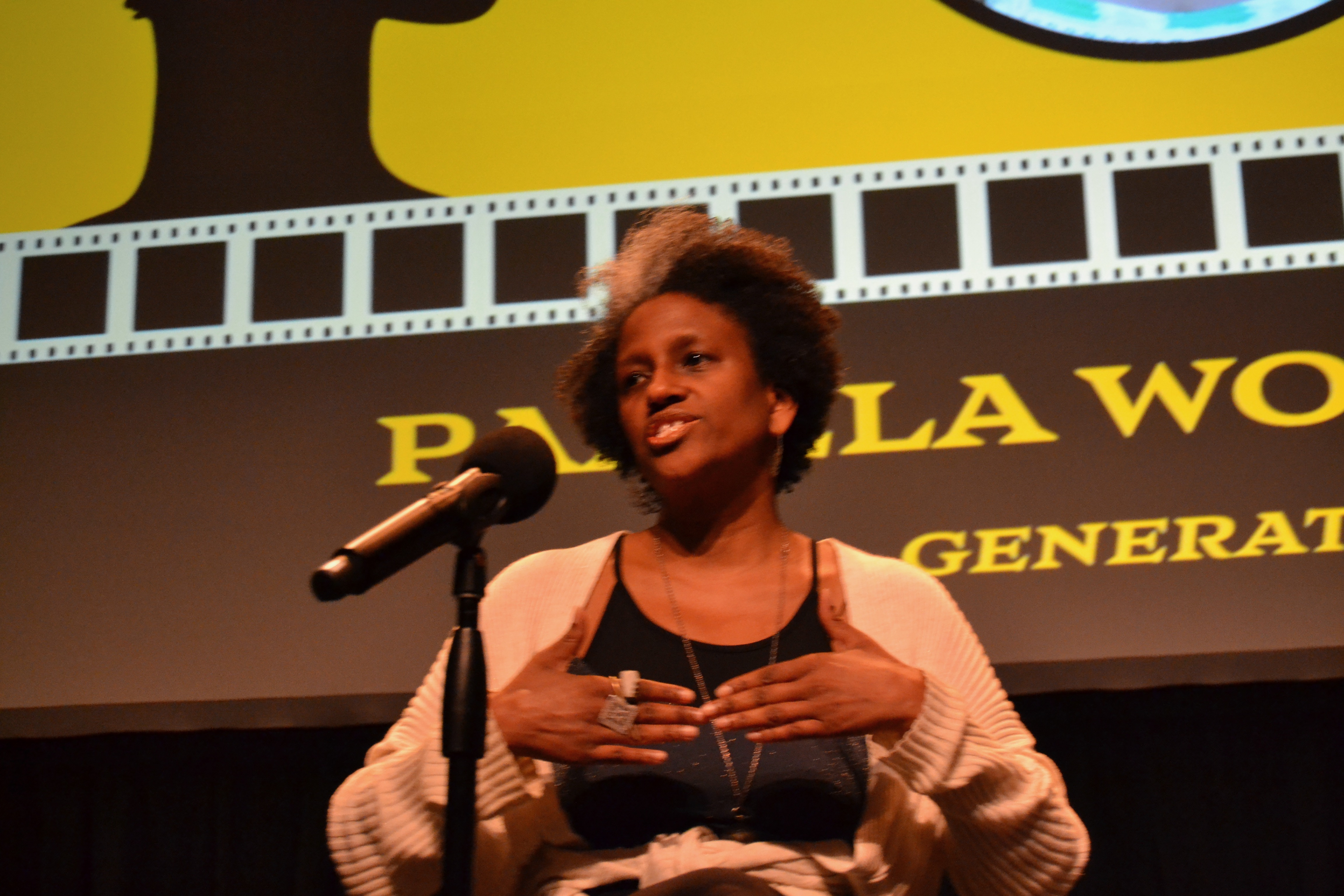 Pamela Woolford being expressive with her hands while speaking in front of a mic in front of a huge screen with parts of the words "PAMELA WOOLFORD, GENERATION" showing on the screen along with part of huge filmstrip graphics.