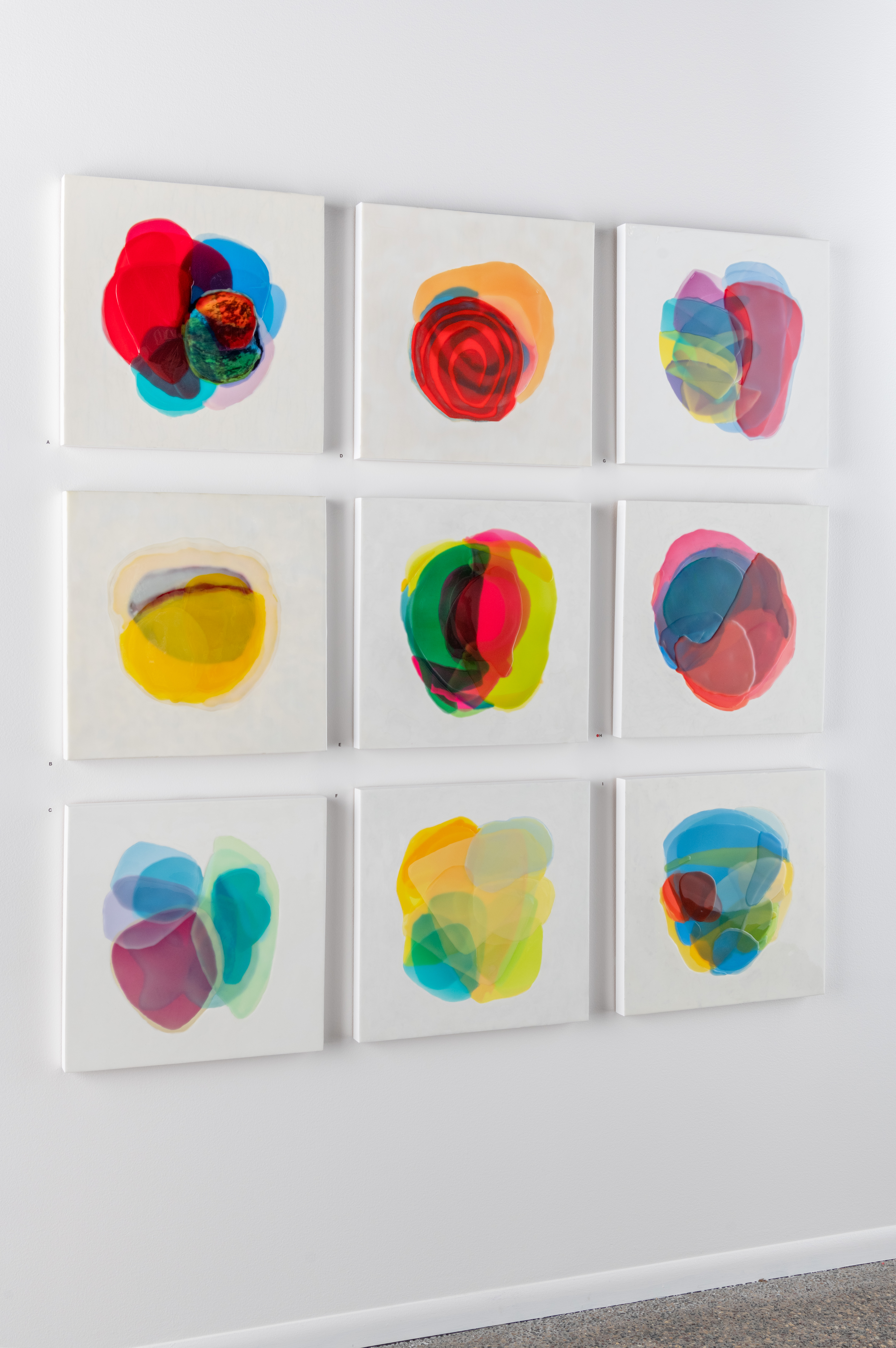 Wall of nine paintings each with colorful abstract forms on white backgrounds by Farida Hughes