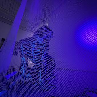 a person wearing a full body suit covered in a glowing nervous system outline crawl an the floor behind a mesh scrim.