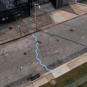 Aerial photo of a public art installation mapping the path of an underground stream. A bright blue wavy line snakes all the ways across a roadway and sidewalks between a grass strip and a black metal fence.