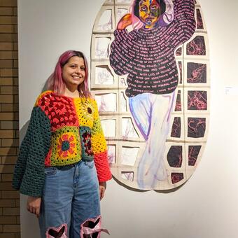 Annika, the artist, stands to the left of her painting of herself, pulling hair away from her face. Words from the AQ are written on her black shirt, and there are gridded cut-outs in the background with threads tangled peeking out