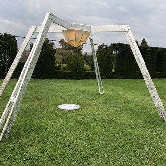 This is a daytime view of the sculpture.A wooden sculpture with four structural diagonal legs laminated with reflective metal foil. The central illuminated lantern contains a projector focused on the ground within the structure, that is rotating and changing in hue.