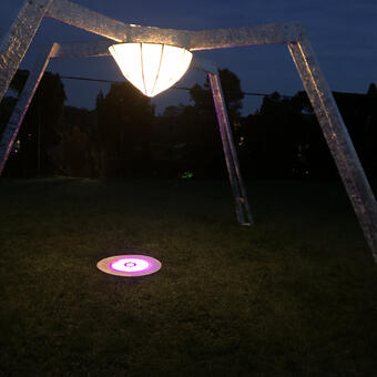 A wooden sculpture with four structural diagonal legs laminated with reflective metal foil. A central illuminated lantern contains a projector focused on the ground within the structure. 