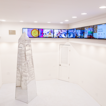 Modern art gallery interior with a sleek white space featuring a metallic rocket-shaped sculpture covered in newspaper clippings at the center. Above, a stretched panoramic display shows a sequence of colorful digital images and text messages, creating an immersive multimedia experience. The gallery's clean lines and bright lighting highlight the contemporary nature of the artwork