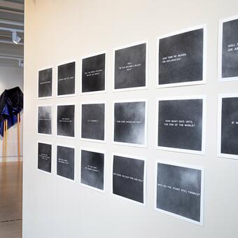 a wall of small graphite drawings in the foreground with gallery installation in the background