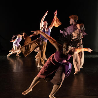 A group of dancers in rich earth tone costumes in dynamic physical movements in space.