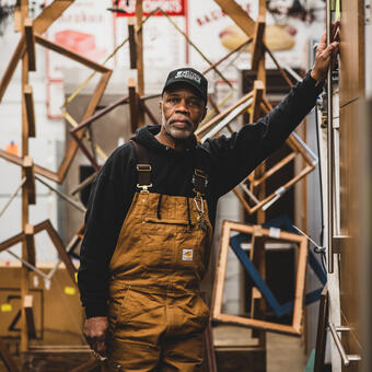 A man in mustard colored overalls poses thoughtfully for the camera with an arm leaning against the wall with discarded wooden picture frames behind him 