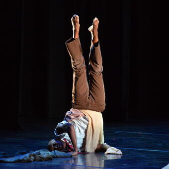 A male dancers suspended in an inversion, balancing on shoulder and side of neck. Dancer is wearing a sleeveless white shirt, red necktie, and chocolate brown pants, bare feet