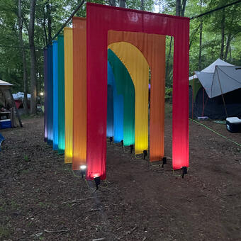 A series of colorful fabric doorways suspended in a forest, with trees and tents visible in the background. 