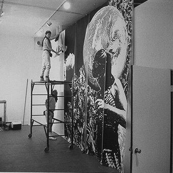 Woman stands on a scaffold to hang 13 ft vellum panels from a large format printer. Thirty-six black and white images fill the entire room. Two bystanders look on.