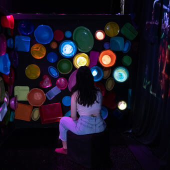 Person sitting in a dark room surrounded by plastic containers with videos projected into 10 containers