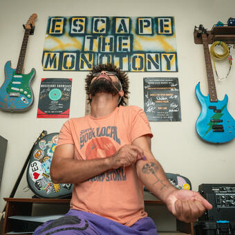 Tavair kneels in front of a wall covered in art that includes 4 guitars and a banner that reads, "Escape the Monotony." He has a guitar string wrapped around his upper arm and the other end is being pulled by his mouth clenched onto it with his head back. At the vein in the middle of that arm he places a guitar pick.