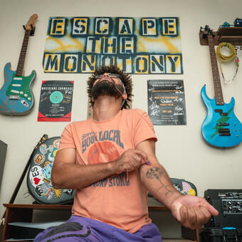 Tavair kneels in front of a wall covered in art that includes 4 guitars and a banner that reads, "Escape the Monotony." He has a guitar string wrapped around his upper arm and the other end is being pulled by his mouth clenched onto it with his head back. At the vein in the middle of that arm he places a guitar pick.