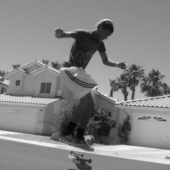 greyscale photograph of skate boarder doing a board flip with houses and palm trees behind him