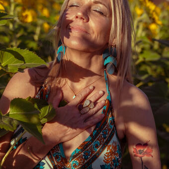 Pam stands in the midst of a sunflower field. Sun and shadow on her face and body with her right hand over her heart. On her left upper arm is a tattoo of a flower. She gazes up to the sky with her eyes closed.