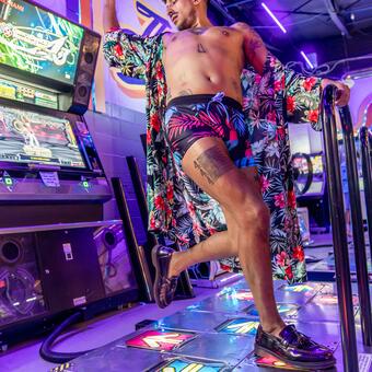 106.7Kris dances on the video game dance dance revolution with his right arm flying behind him and his left leg feeling back for the dance floor. He wears a brightly colored robe and short shots revealing several tattoos. The point of focus is the 444 tattoo on his upper right thigh.