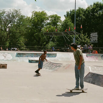 Two skateboarder, one in a greenshirt in the forefront and a shirtless African American male in the background, skating in a graffiti covered park
