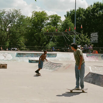 Two skateboarder, one in a greenshirt in the forefront and a shirtless African American male in the background, skating in a graffiti covered park