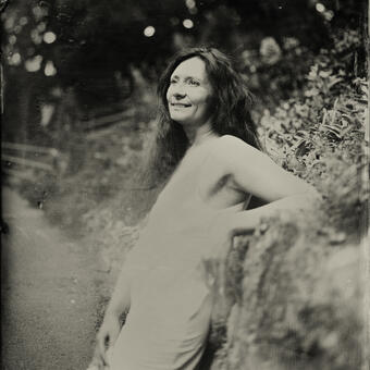 A 3/4 view of a woman's body; she is leaning on the rock and smiling, looking away.  Her hair is black and long, and she's wearing a white dress. Wet plate collodion