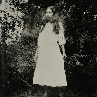 A wet plate collodion photograph of a woman, full body; wearing white dress.  There is slight movement in the body due to slow exposure