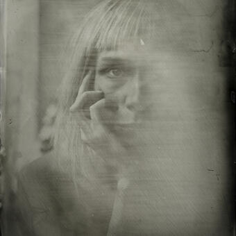 A close up photograph of a woman, made with wet plate collodion process.  The woman is looking at the viewer, and her hand is touching her face.  The image has collodion artifacts due to fogging, and the face is barely visible though the fog. 