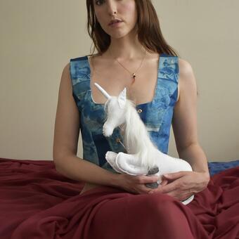 A white woman with long brown hair sits on a bed in a vest made of cyanotypes of Italian renaissance paintings featuring unicorns. She holds a white fabric unicorn in her lap and has burgundy chiffon fabric draped over her lower half. 