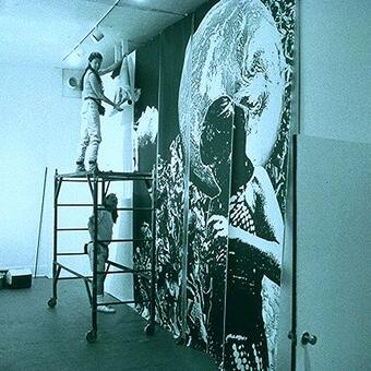 An artist stands on a scaffold installing thirteen foot high panels along a wall