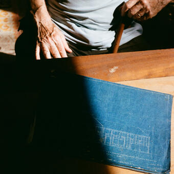 Photo of man's hands holding cane with blueprint on the table in front of him, half in shadow.