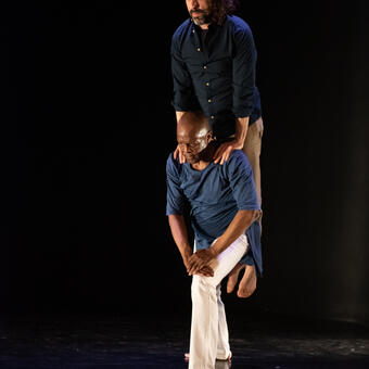 Two males, one in white pants and blue tunic, the other in tan pants and dark blue shirt. One male is in a forward left leg lunge and the other is on his shoulders extending his body over his head (like a totem pole statue). Both are looking out towards a horizon.