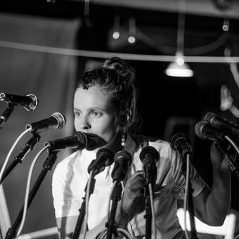 A woman stands in front of 12 microphones, speaking into one