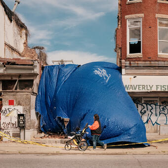 A woman in an Orioles jacket pushes a stroller past a collapsed building covered by a massive, weathered blue tarp. The remnants of the Waverly Fish Market and a decayed storefront with graffiti and torn posters frame the scene, caution tape marking the dereliction.