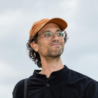 Bruce Willen, looking slightly up and to the right, wearing glasses, and orange baseball hat and black shirt, against a background of sky