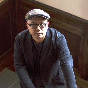 Lysley Tenorio, clad in hat, glasses, and navy blazer, standing in stairwell, looking directly at viewer