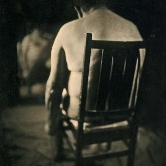 Image of person sitting in a chair without clothes facing away and reflection of the person in a mirror.