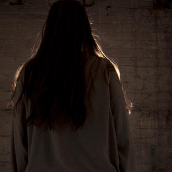Woman back turned to viewer facing a dark grimey grey wall. Her figure highlighted/outlined in light.