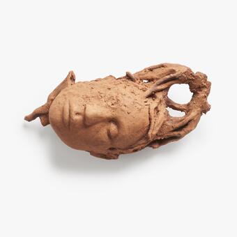 A fragmented ceramic head is positioned horizontally. The hair on the portrait is abstracted and extends into root structures. The sculpture is textured like soil and is the color of red clay.