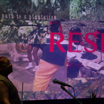 Muse stands in fron of screen, text reads 'RESIST' in bold read overlaid with colorful videos. 