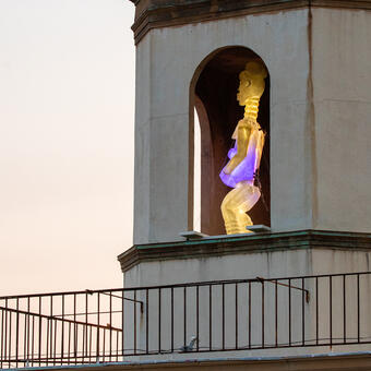Illuminated translucent sculpture of a pregnant standing figure installed inside a historic bell tower at dusk, glowing with blue and violet light above a city intersection.
