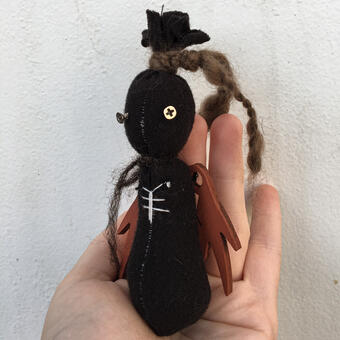 A small black stuffed creature with screws for eyes, with strings wrapped around it. It sits in the hand of a person. 