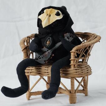 A small black hooded stuffed creature with a yellow mask with a tiny wooden lute like instrument sits on a wicker chair. 