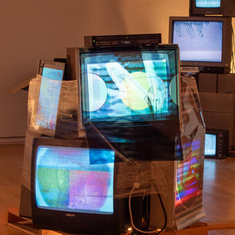 A Pallet of Living, Injured, and Dead Electronics, LCDs, CRTs, sits on an industrial moving Pallet in the middle of the gallery where a viewer can walk 360 degrees around the video sculpture