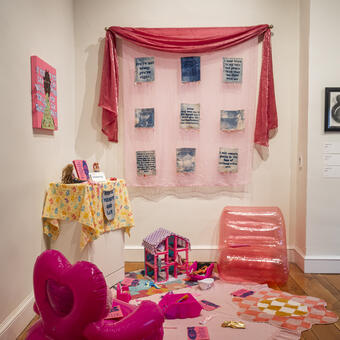 A corner of the room with a painting, fabric wall art, and a play area with dolls and a pink doll house and plastic chairs. 