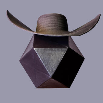 Abstract portrait of a purple geometric form made from papermache wearing a wide brimmed woman's hat folded to look like wings. Photographed in studio against a lilac colored background.