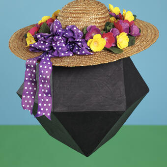 Abstract portrait of a black geometric form made from papermache wearing a woman's gardening hat with ribbon and flowers. Photographed in studio against a background styled to look like an abstract representation of a sumemr day.
